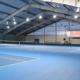 Kryty kort tenisowy z niebieską nawierzchnią, widoczne siatki, oświetlenie sufitowe i ekran projekcyjny w tle.
