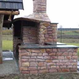 Murowany grill ogrodowy z kamienia naturalnego, z blatem z ciemnego kamienia, zadaszony drewnianą konstrukcją, na tle trawnika i ogrodzenia.