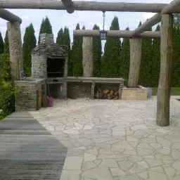 Ogród z kamienną podłogą, grillem i zadaszeniem z drewnianych bali, otoczony tujami.