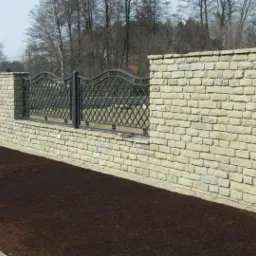 Kamienny mur z kutym, metalowym ogrodzeniem w tle, świeżo zasiana ziemia wzdłuż muru, widok z poziomu gruntu.