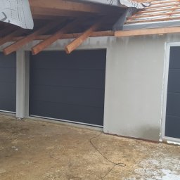 PHTU CONTROL - Dwie grafitowe bramy garażowe segmentowe z ościeżnicami i osobne grafitowe drzwi wejściowe zamontowane w surowym budynku z widoczną konstrukcją dachu i niewielkimi śladami śniegu na ziemi.