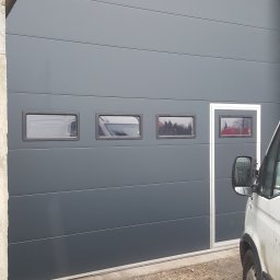 PHTU CONTROL - Szara segmentowa brama garażowa z czterema małymi okienkami, widoczny fragment białego samochodu dostawczego po prawej stronie, szare tło betonowego budynku.