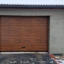 PHTU CONTROL - Brązowa brama garażowa segmentowa z poziomym wzorem drewna, zamontowana w surowym, szarym garażu z widocznymi śladami po tynkowaniu, na tle ośnieżonego podjazdu i żółtego budynku.