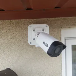 Zewnętrzna kamera monitoringu BCS zamontowana na elewacji budynku, widoczna obok okna, pod brązowym okapem dachu.