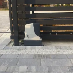 Szara, pozioma brama przesuwna z czarnymi profilami i napędem Nice Robus, zamontowana na szarej kostce brukowej, widok z bliska na mechanizm i fragment podjazdu.