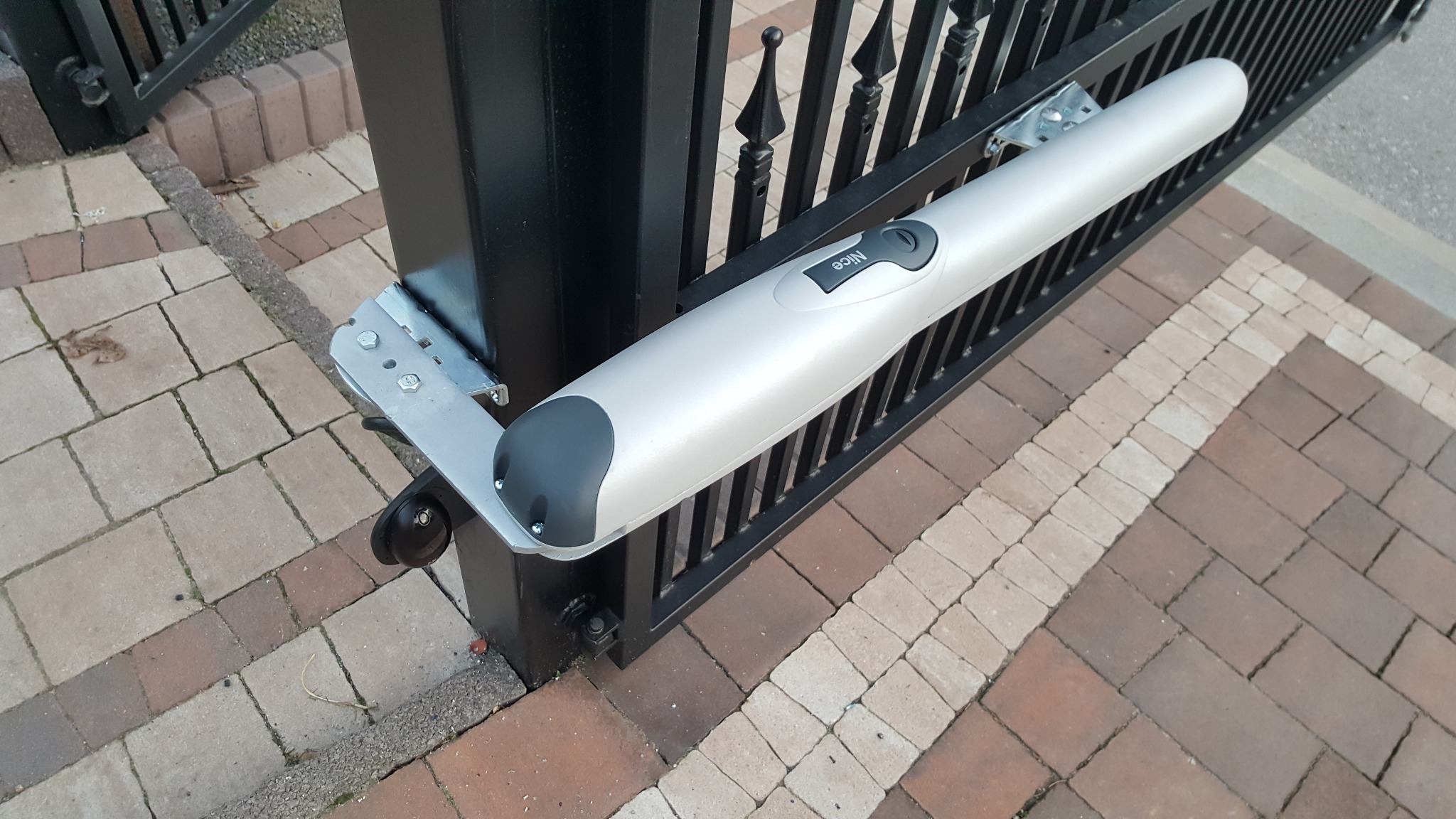 Siłownik NICE WINGO 2024 zamontowany na czarnej, metalowej bramie, widoczny z boku na tle kostki brukowej. Widoczne śruby mocujące i logo producenta.