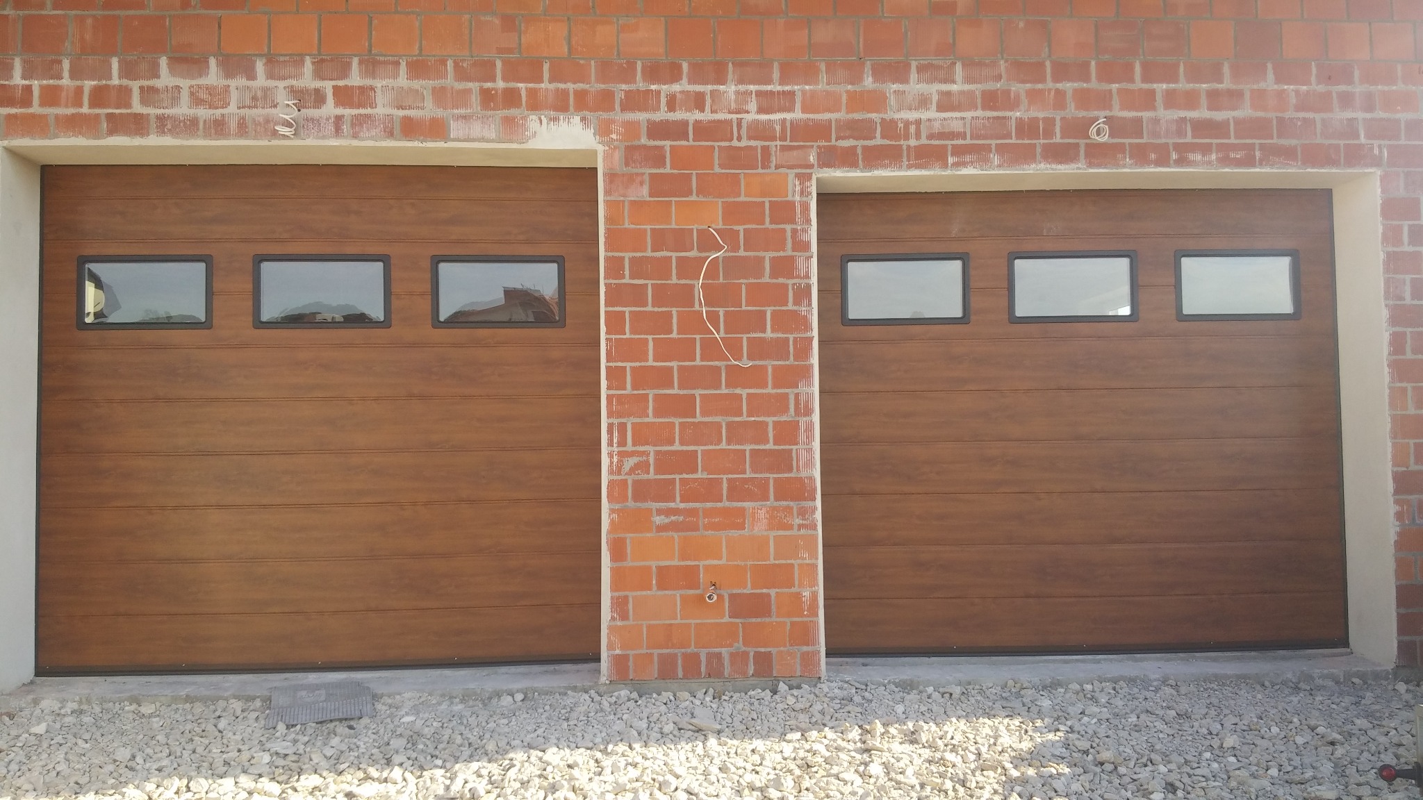 Dwie brązowe segmentowe bramy garażowe z trzema prostokątnymi okienkami, zamontowane w surowej, ceglanej fasadzie budynku w trakcie budowy, widoczne kamienie i przewody.
