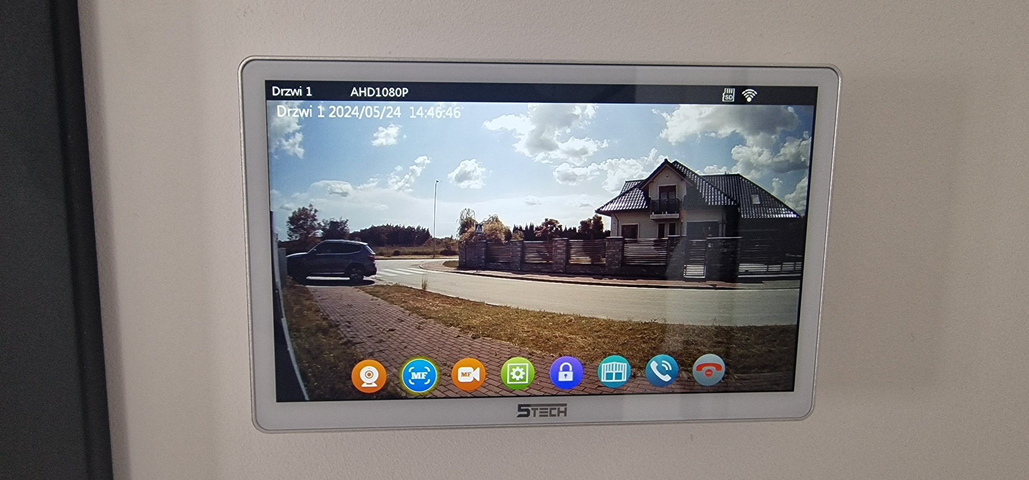 Ekran wideodomofonu z podglądem na posesję: dom z ciemnym dachem i ogrodzeniem, samochód przy ulicy, data i godzina nagrania w lewym górnym rogu.