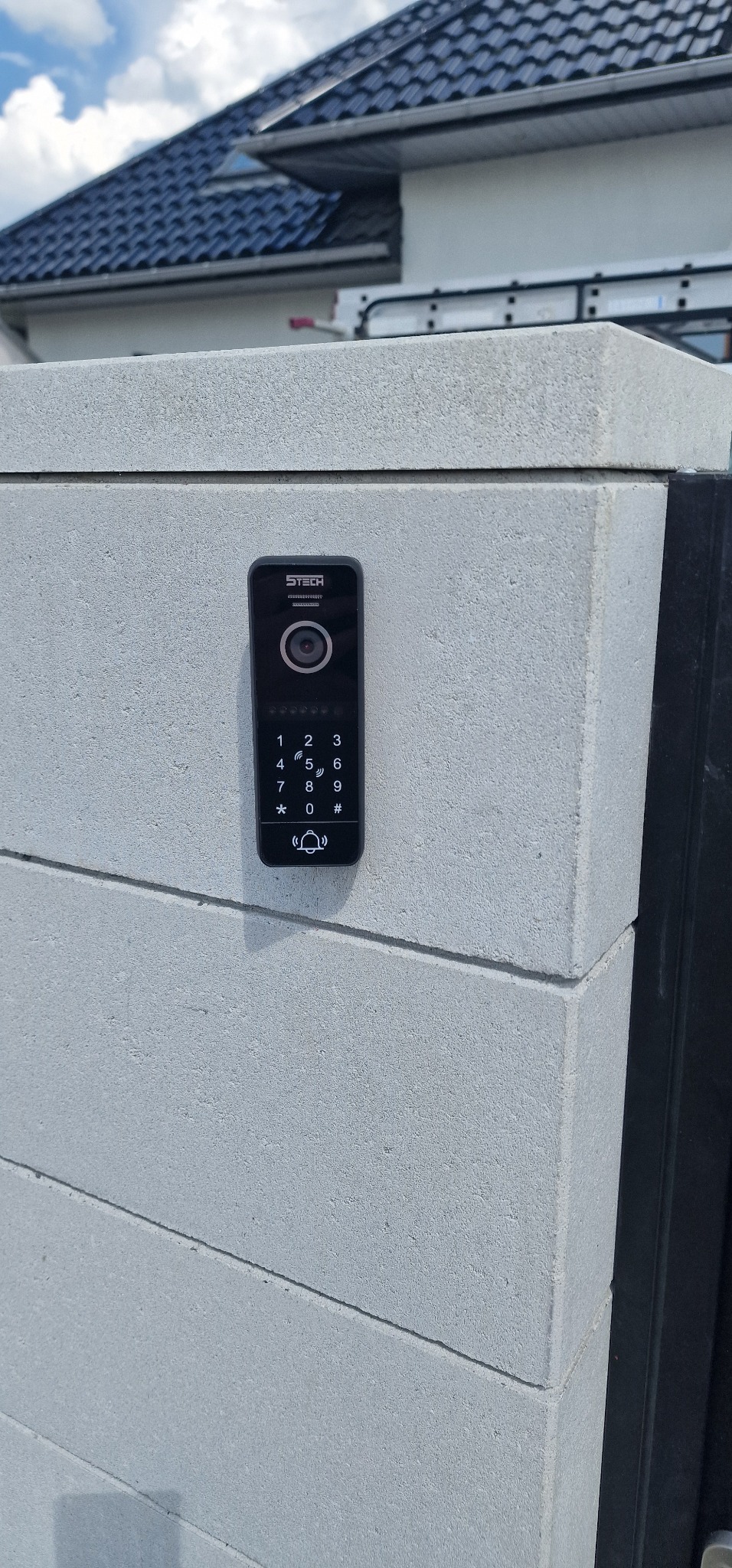 Czarny wideodomofon z klawiaturą numeryczną zamontowany na szarym, betonowym słupku ogrodzeniowym, widoczny fragment dachu domu.