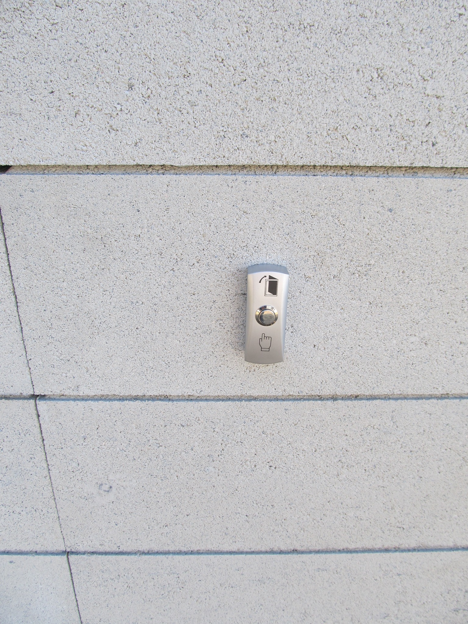 Srebrny przycisk domofonu z symbolem otwierania drzwi i dłoni wskazującej na ścianie z szarego betonu komórkowego.