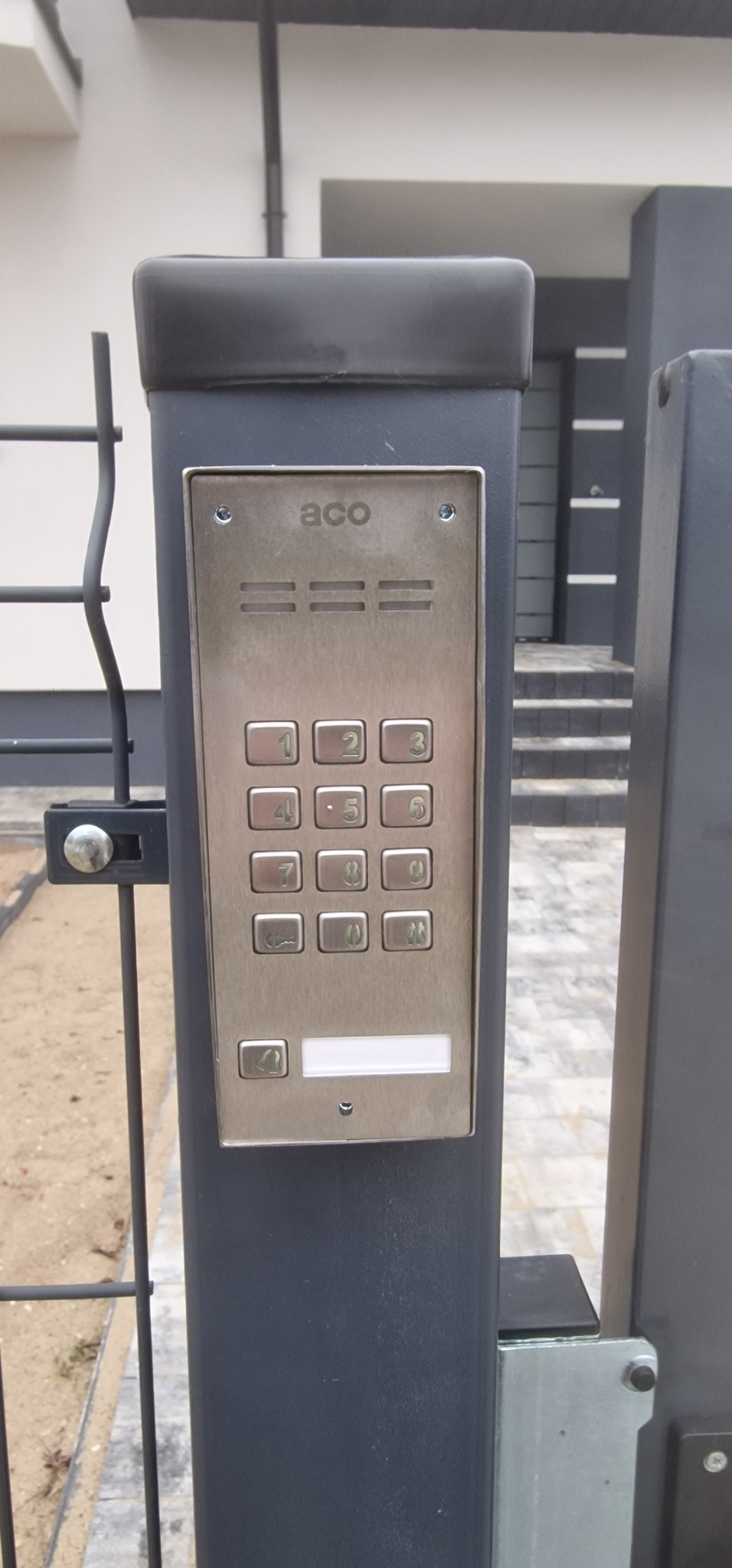Panel domofonu ACO z klawiaturą numeryczną zamontowany na grafitowym słupku ogrodzenia, widoczne schody w tle.