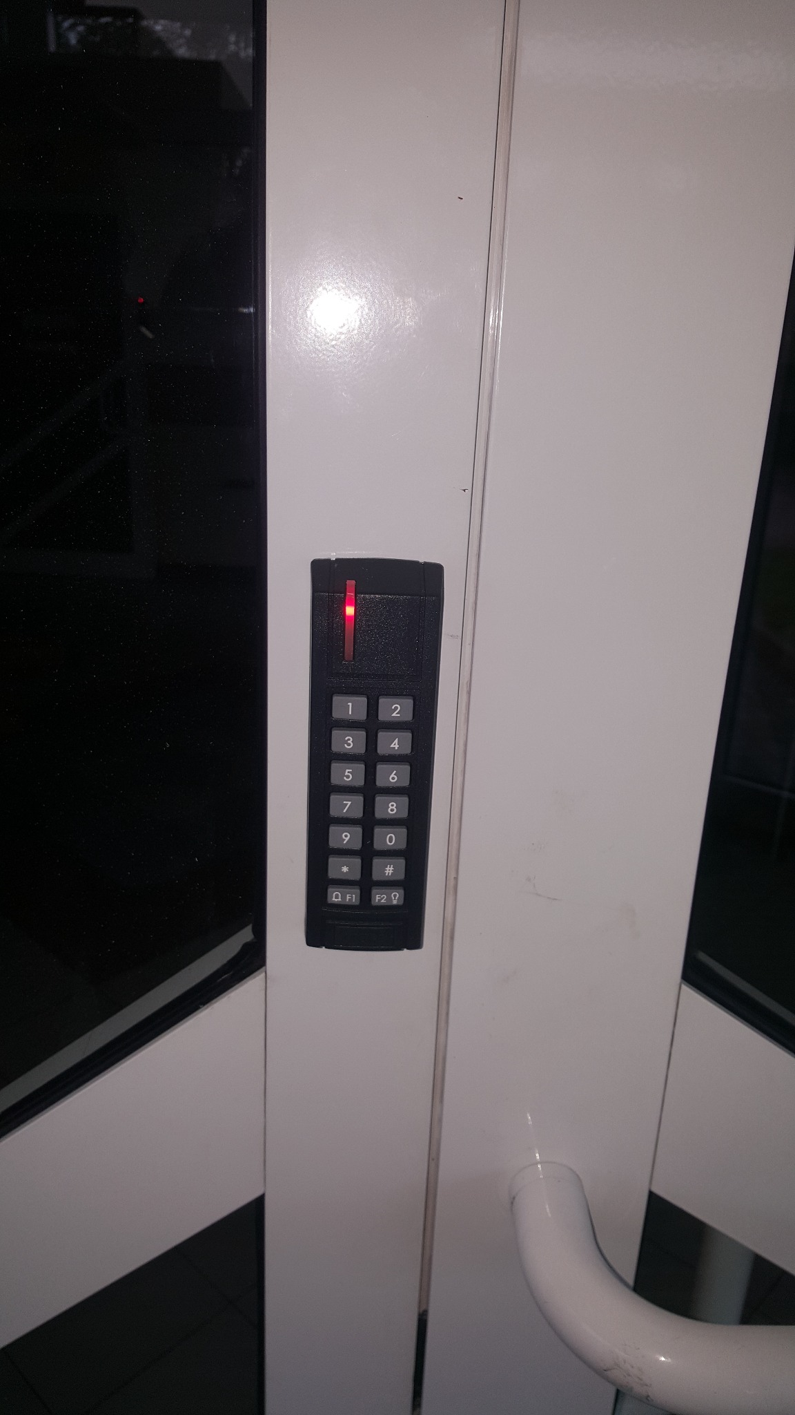 Klawiatura numeryczna kontroli dostępu z czerwoną diodą LED zamontowana na białych drzwiach z czarnymi szybami.