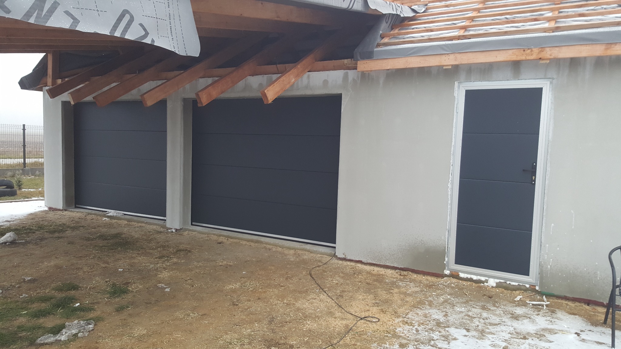 Dwie grafitowe bramy garażowe segmentowe z ościeżnicami i osobne grafitowe drzwi wejściowe zamontowane w surowym budynku z widoczną konstrukcją dachu i niewielkimi śladami śniegu na ziemi.