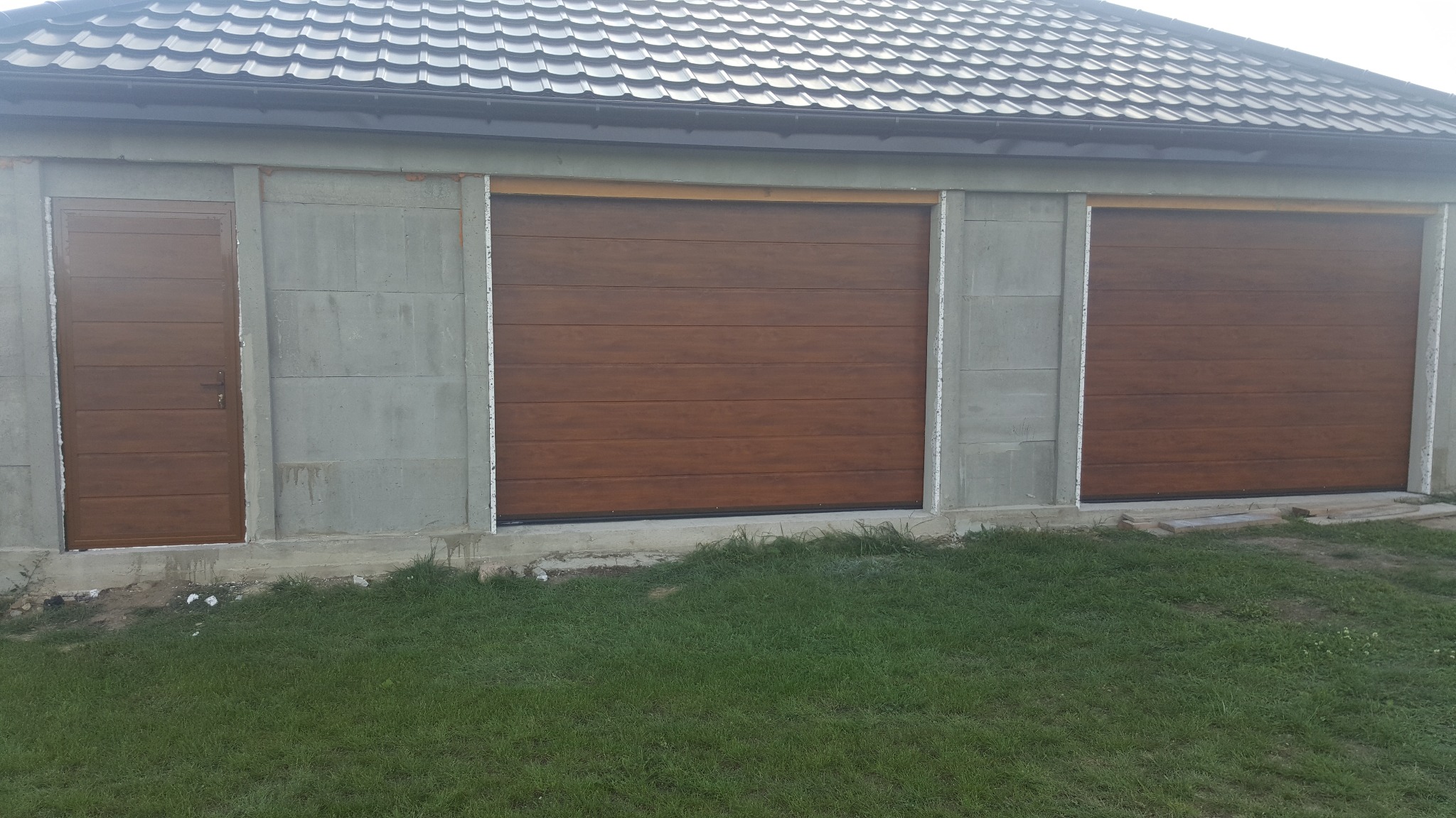 Dwie brązowe bramy garażowe segmentowe oraz drzwi wejściowe o identycznym wzorze, zamontowane w surowym, betonowym budynku z szarą dachówką.