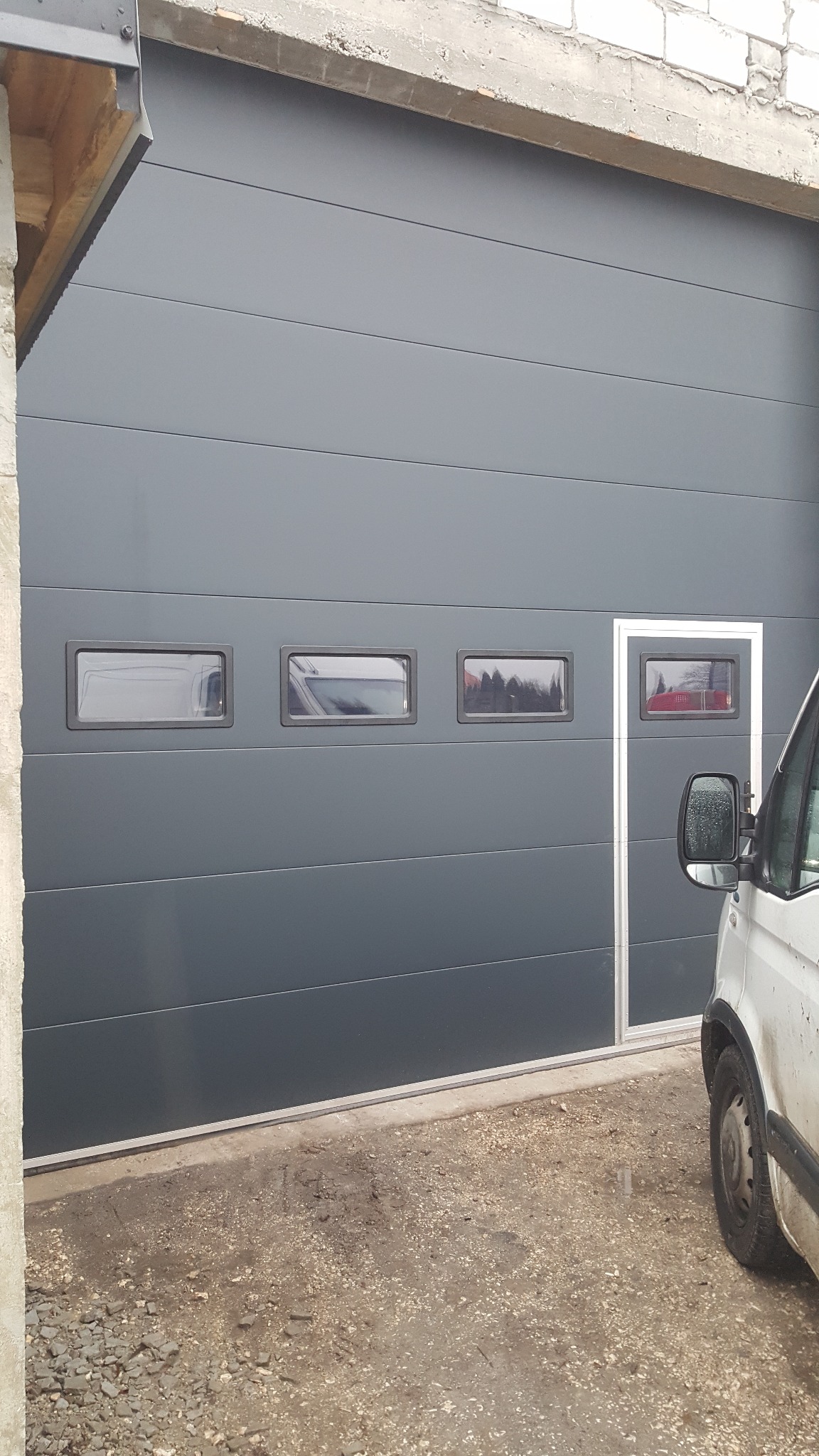 Szara segmentowa brama garażowa z czterema małymi okienkami, widoczny fragment białego samochodu dostawczego po prawej stronie, szare tło betonowego budynku.