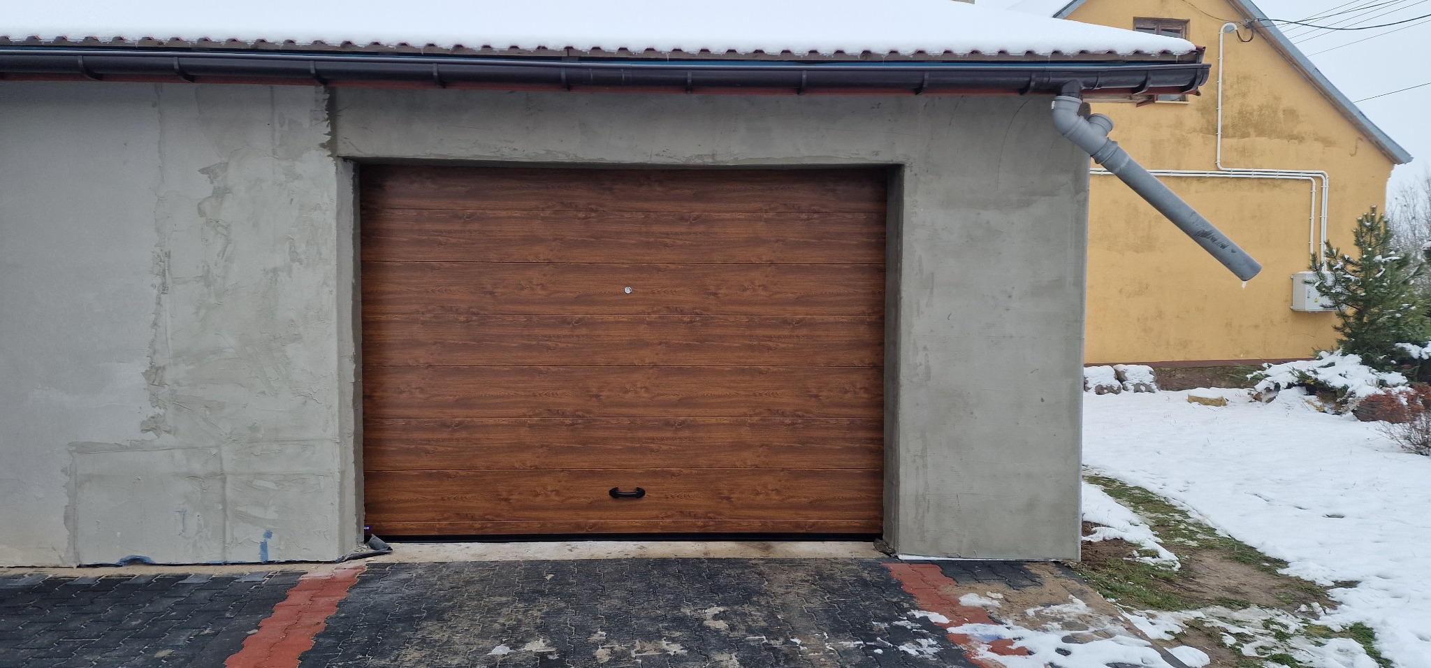 Brązowa brama garażowa segmentowa z poziomym wzorem drewna, zamontowana w surowym, szarym garażu z widocznymi śladami po tynkowaniu, na tle ośnieżonego podjazdu i żółtego budynku.