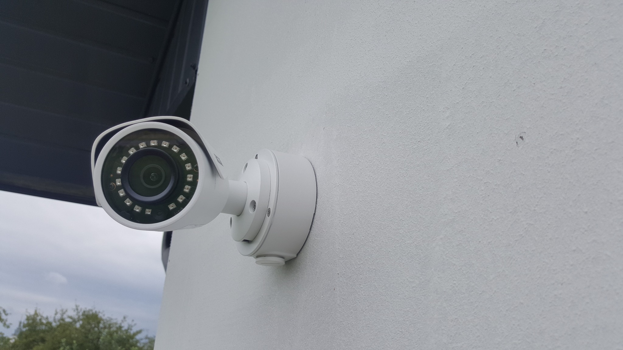 Biała kamera monitoringu zewnętrznego z diodami IR zamontowana na chropowatej ścianie budynku, widoczne fragmenty dachu i zieleni w tle.