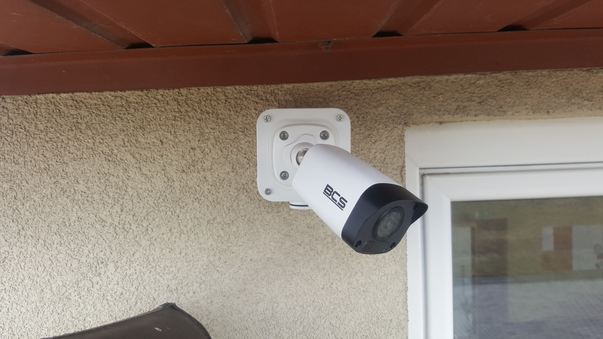 Zewnętrzna kamera monitoringu BCS zamontowana na elewacji budynku, widoczna obok okna, pod brązowym okapem dachu.