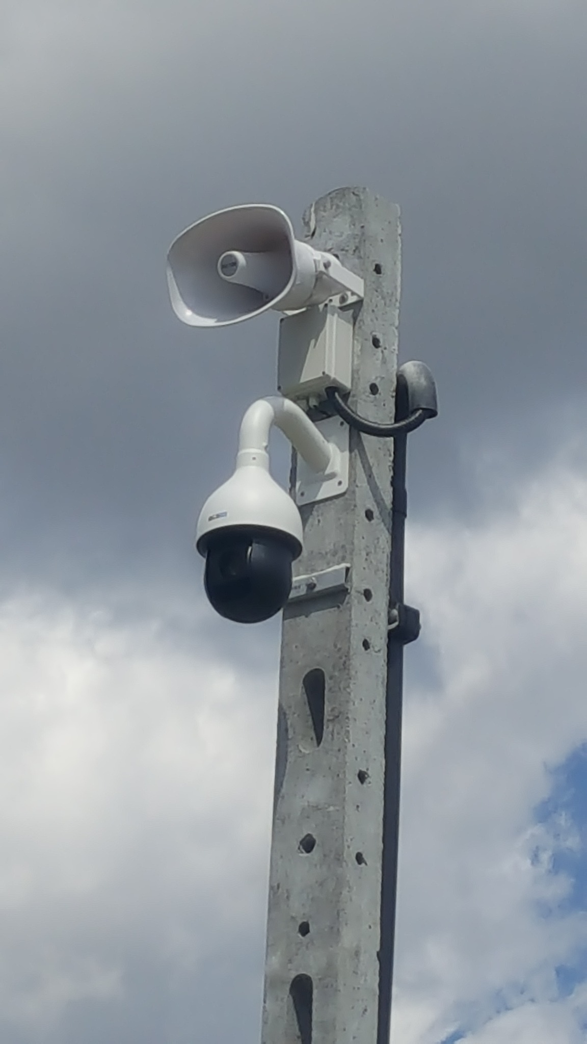 Kamera PTZ i głośnik tubowy zamontowane na betonowym słupie na tle pochmurnego nieba.