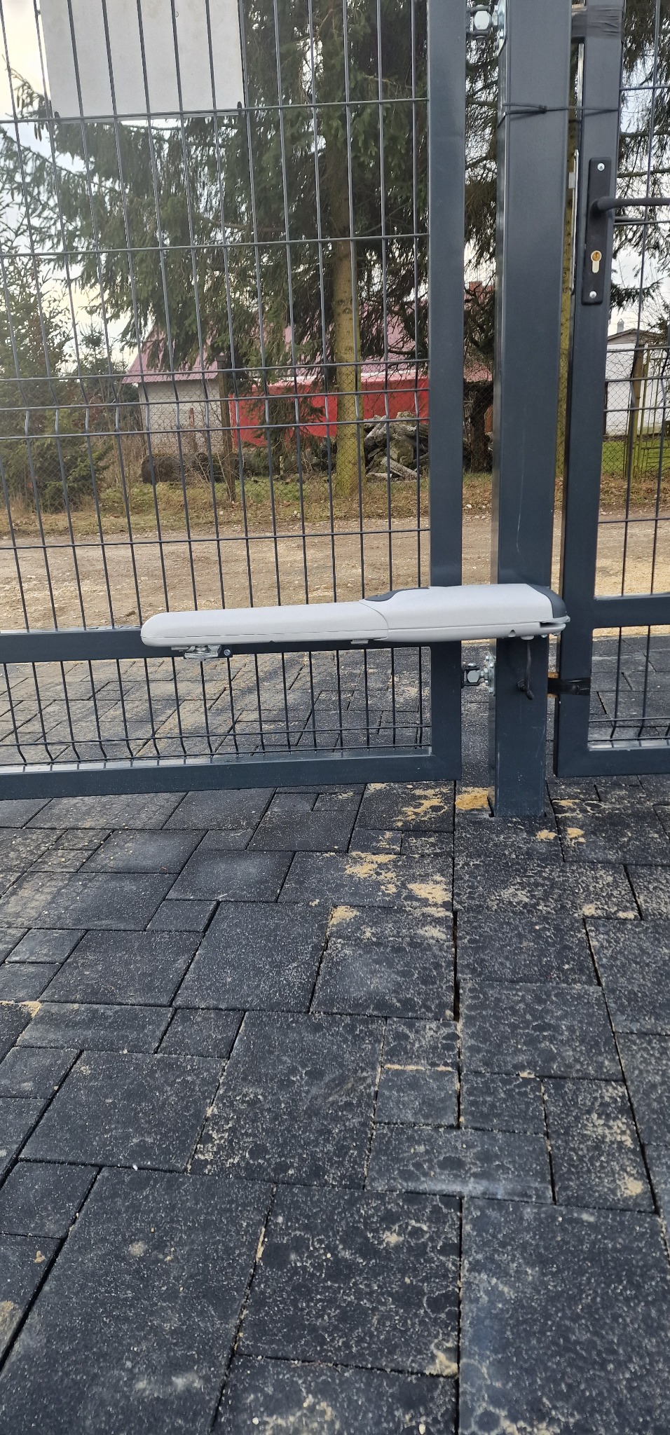 Szara brama z siatki z zamontowanym białym siłownikiem automatyki, widok na czarną kostkę brukową i dom w tle.