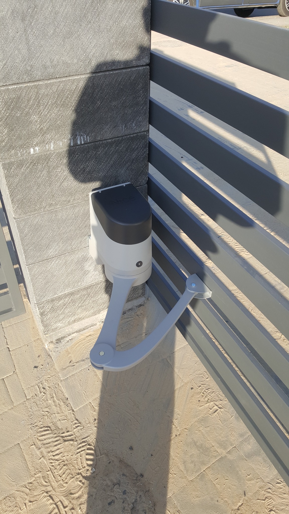 Szary napęd do bramy skrzydłowej zamontowany na słupku z bloczków betonowych, widoczne ślady butów na piaskowej nawierzchni.