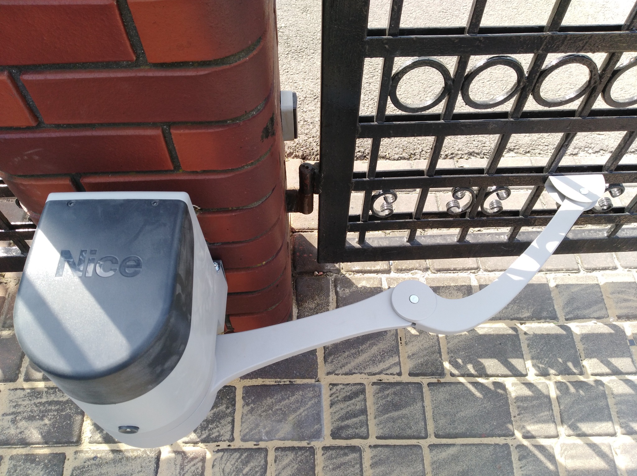 Napęd bramy skrzydłowej marki Nice zamontowany na murowanym słupku, widoczna metalowa brama z ozdobnymi elementami.