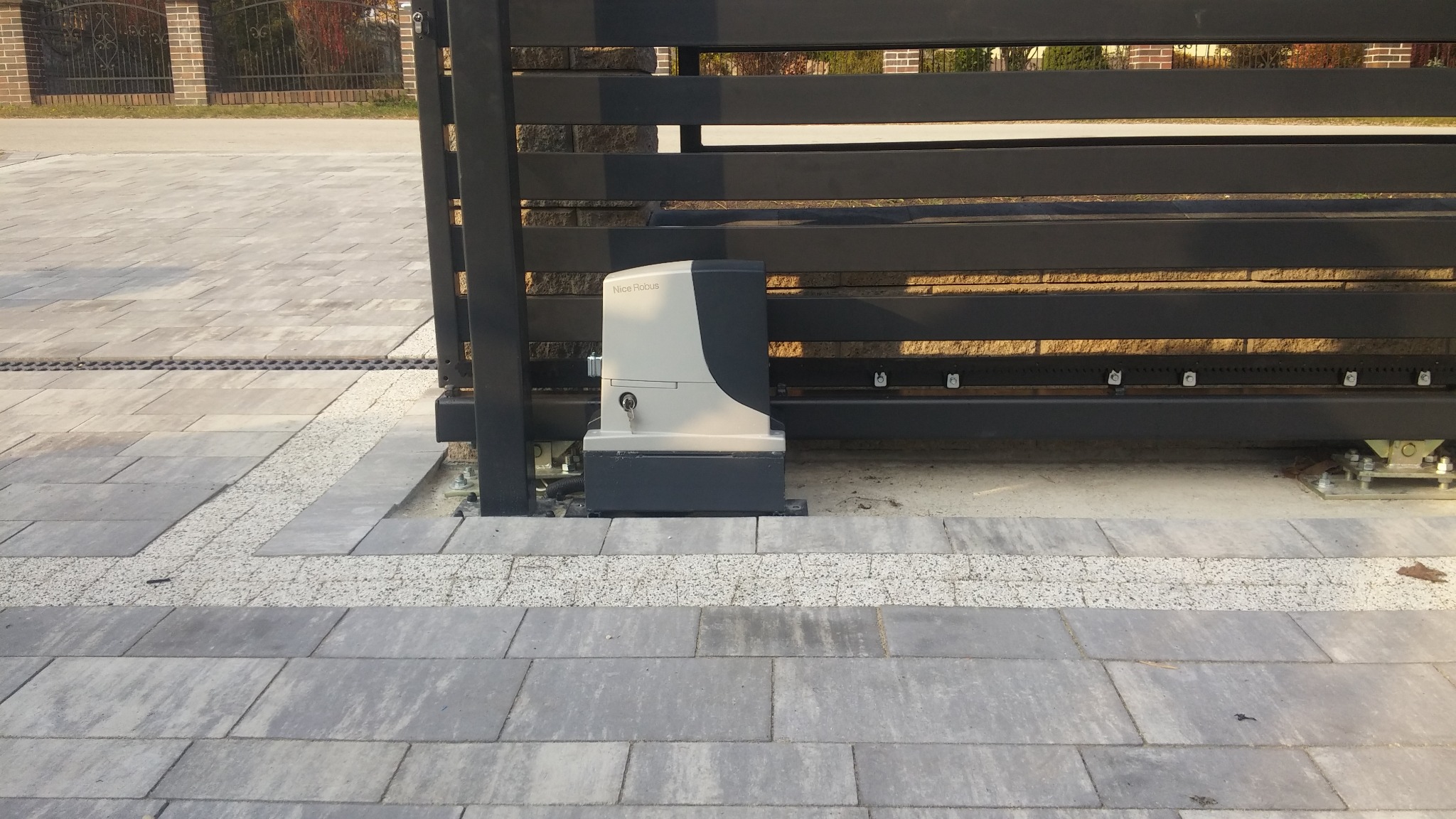 Szara, pozioma brama przesuwna z czarnymi profilami i napędem Nice Robus, zamontowana na szarej kostce brukowej, widok z bliska na mechanizm i fragment podjazdu.