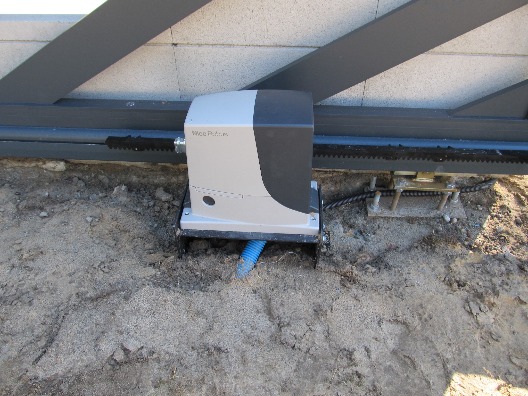 Szary napęd do bramy przesuwnej marki Nice Robus zamontowany na ziemi obok zębatki. Widoczna niebieska rura osłonowa kabli.
