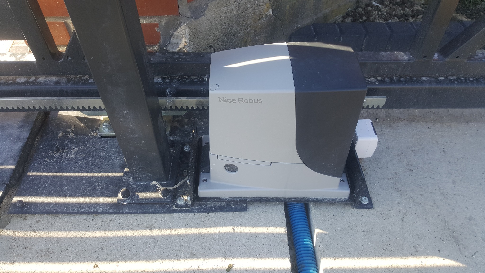 Szary napęd Nice Robus zamontowany do czarnej bramy przesuwnej z zębatą listwą, widoczna niebieska rura kablowa w betonie