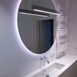 Nowoczesna łazienka z owalnym lustrem podświetlanym LED, białą umywalką wpuszczaną w blat i grzejnikiem drabinkowym w tle.