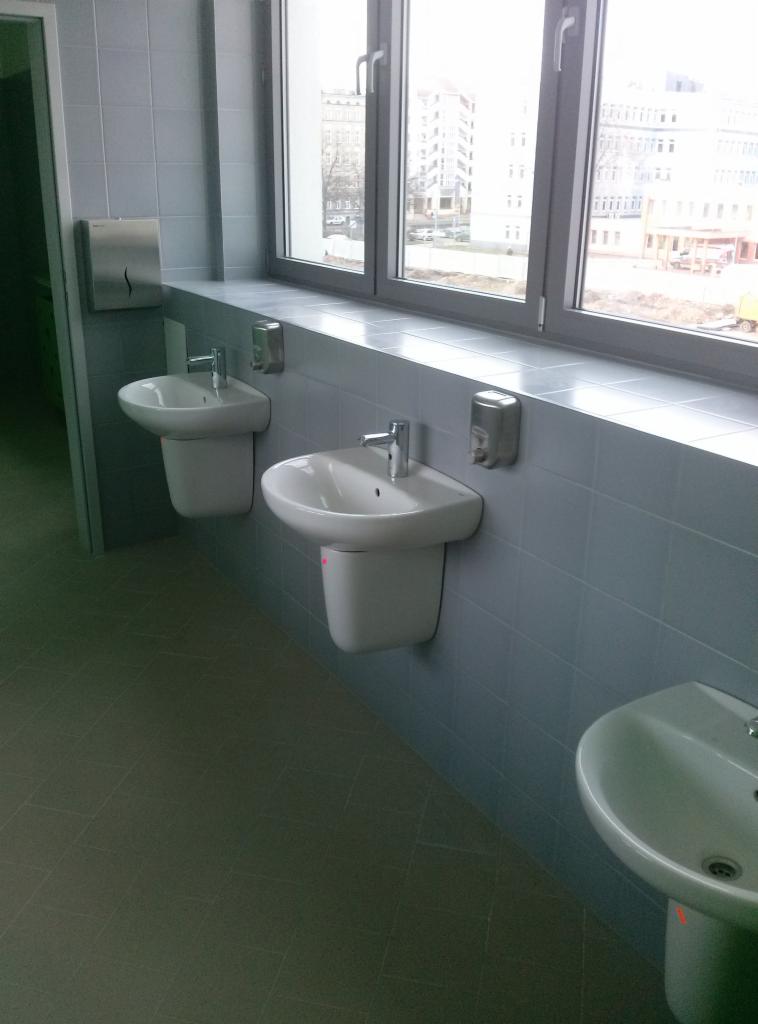 Umywalki zamontowane w rzędzie w łazience szkolnej, widoczne dozowniki mydła i okno z widokiem na budynek szkolny.