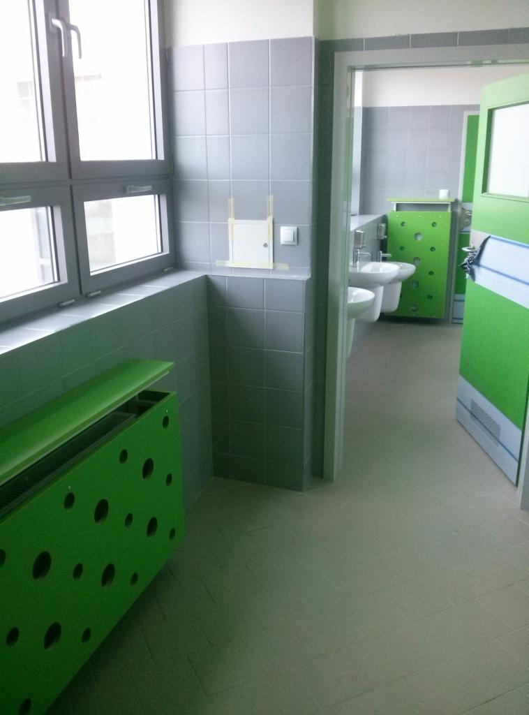 Szara, kwadratowa glazura ścienna w publicznej toalecie szkolnej, z widoczną zieloną obudową grzejnika z okrągłymi otworami i fragmentem instalacji przygotowanej do podłączenia.