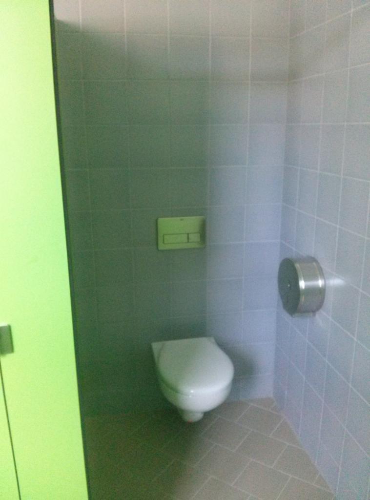 Wnętrze toalety w szkole SP 84 we Wrocławiu, wykończone szarą glazurą z białą fugą, z podwieszaną miską WC i metalowym podajnikiem na papier toaletowy.