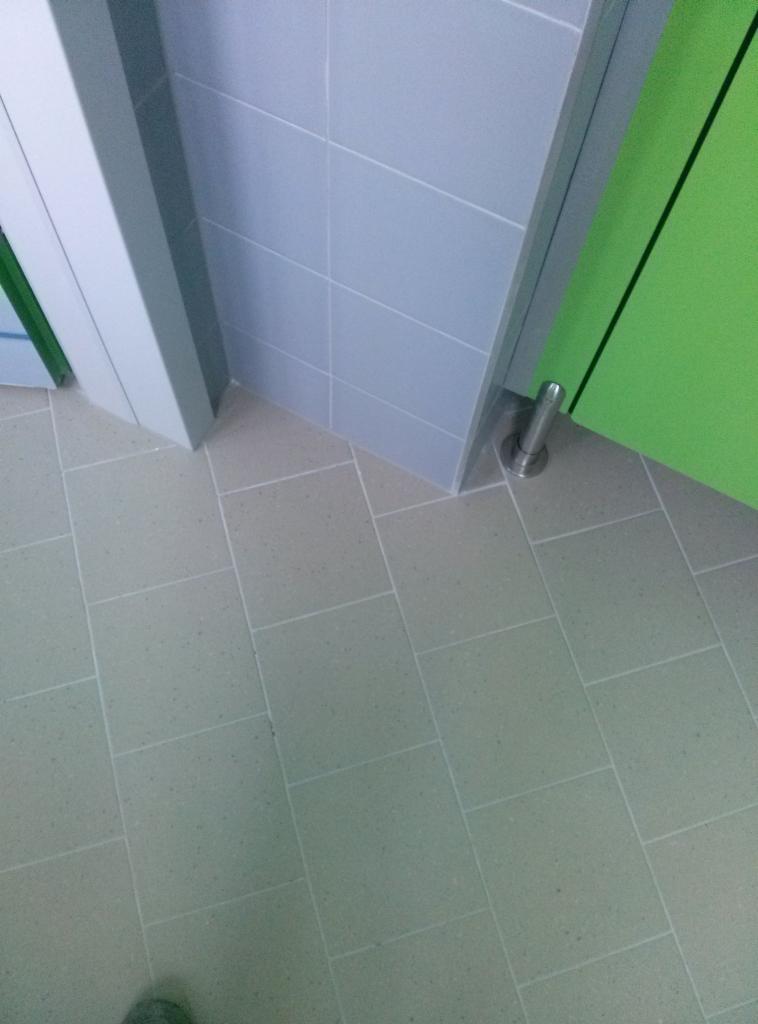 Szare, kwadratowe płytki podłogowe i ścienne w publicznej toalecie, widoczna dolna część zielonych drzwi kabiny oraz metalowy ogranicznik drzwi.