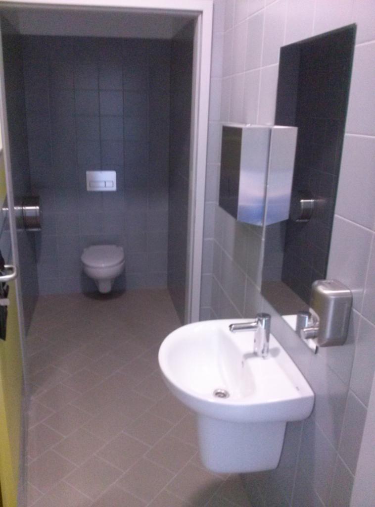 Wykończona łazienka w odcieniach szarości z białą umywalką i toaletą, widoczne dozowniki mydła i papieru, płytki na ścianach i podłodze ułożone w regularny wzór.