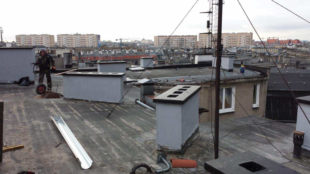 Praca na dachu: dekarz z taczką, świeżo pomalowane kominy, rynna na płaskim dachu z widokiem na miasto i budynki w tle, przewody elektryczne.