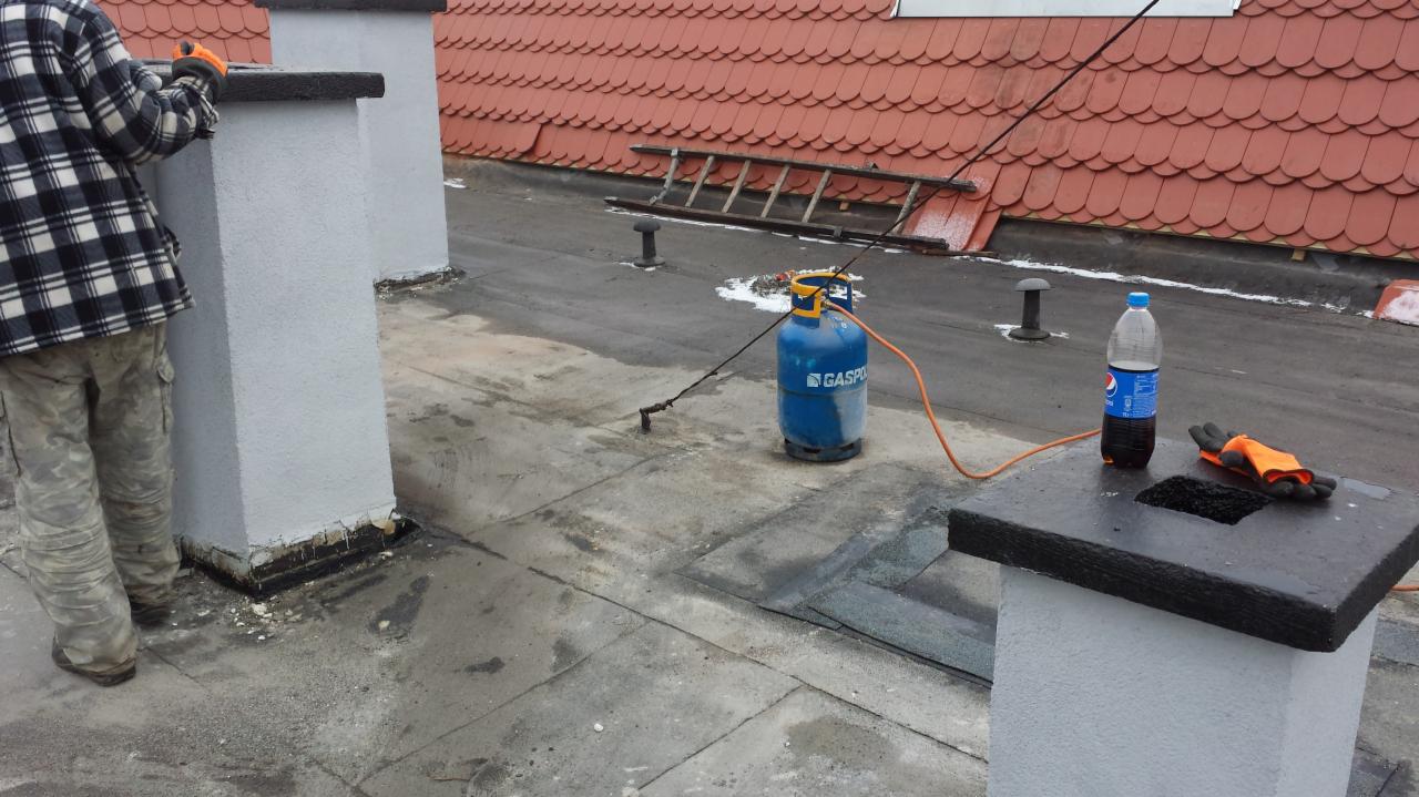 Praca na dachu: dekarz w kratkowanej koszuli przy kominie, butla gazowa, drabina oparta o czerwoną dachówkę, butelka napoju i rękawice. Widok z góry na płaski dach.