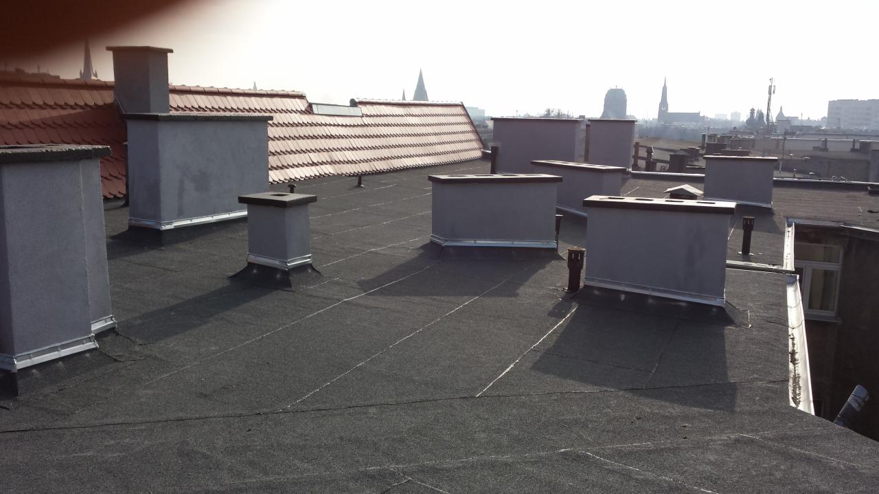 Czarny, płaski dach z kilkoma kominami i fragmentem dachu z czerwonej dachówki w tle. Widok na miasto w tle. Oświetlenie słoneczne, lekka mgła.