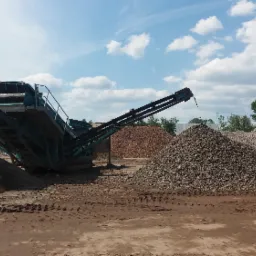 Przesiewacz mobilny sortuje materiał na trzy pryzmy o różnej frakcji, widoczne ślady opon na ziemi, błękitne niebo z obłokami w tle.