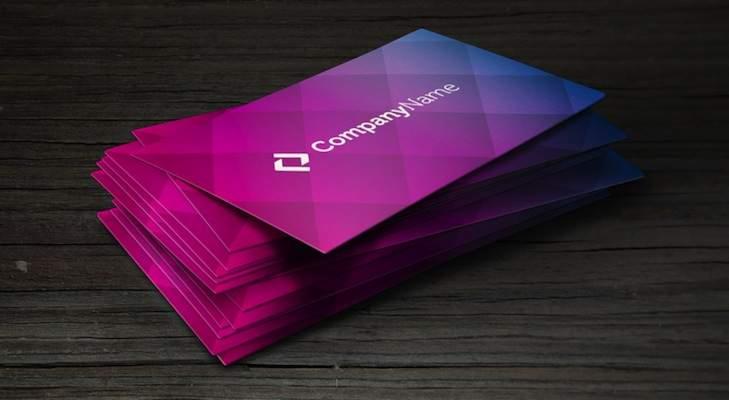 Stos wizytówek z gradientem kolorów od niebieskiego do różowego, leżący na ciemnym, drewnianym tle.