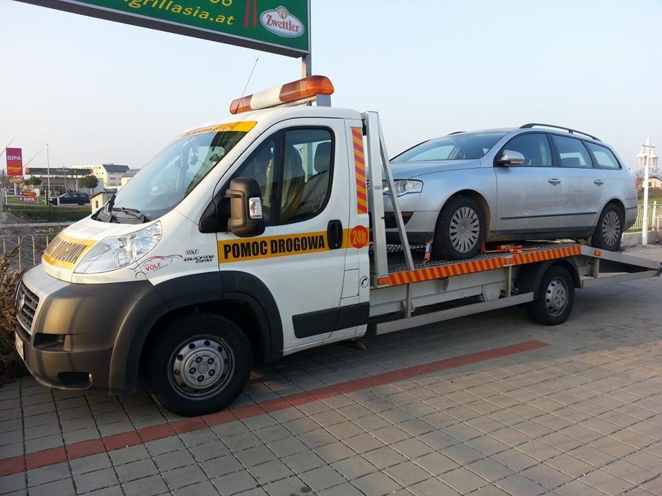 Laweta z srebrnym samochodem osobowym na platformie, zaparkowana na kostce brukowej. Na pojeździe napis 'Pomoc Drogowa 24h'.
