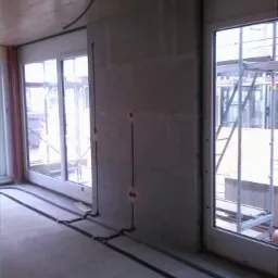 Surowy stan wnętrza budynku w trakcie prac instalacyjnych: widoczne przewody elektryczne prowadzone po podłodze i w ścianach z otworami pod gniazdka, okna z widokiem na rusztowania.