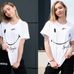Uśmiechnięta blondynka w białym t-shircie z nadrukiem uśmiechu i napisem 'Smile', ubrana w czarne spodnie dresowe z logo 'UBR.CREW', pozuje na tle szarej ściany i fragmentu budynku.