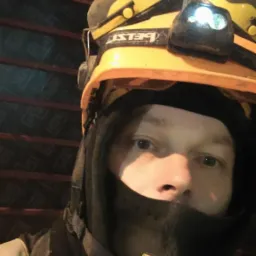 Autoportret pracownika budowlanego w kasku z lampą czołową na tle szalunków. Widoczne elementy rusztowania i zabezpieczenia odzieży roboczej.