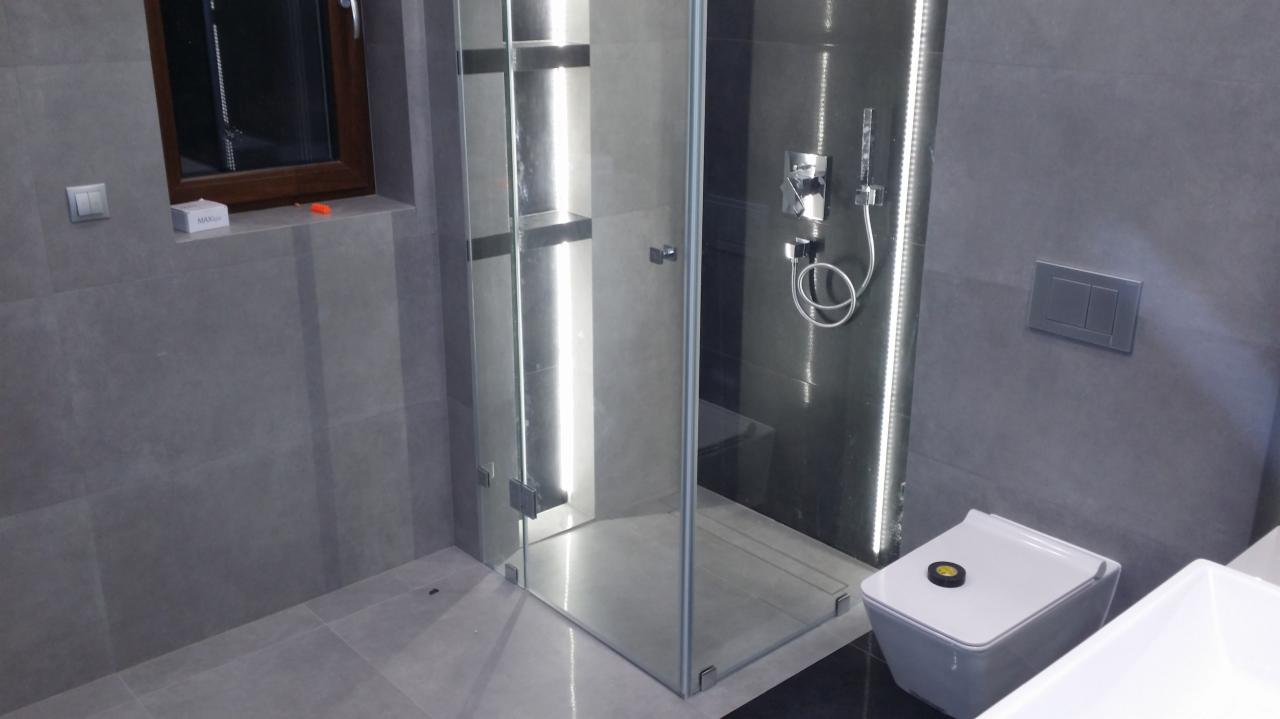 Nowoczesna łazienka z szarymi płytkami, przeszkloną kabiną prysznicową z półkami i deszczownicą, podwieszaną toaletą z czarnym elementem na górze oraz drewnianym oknem z białymi kosmetykami...