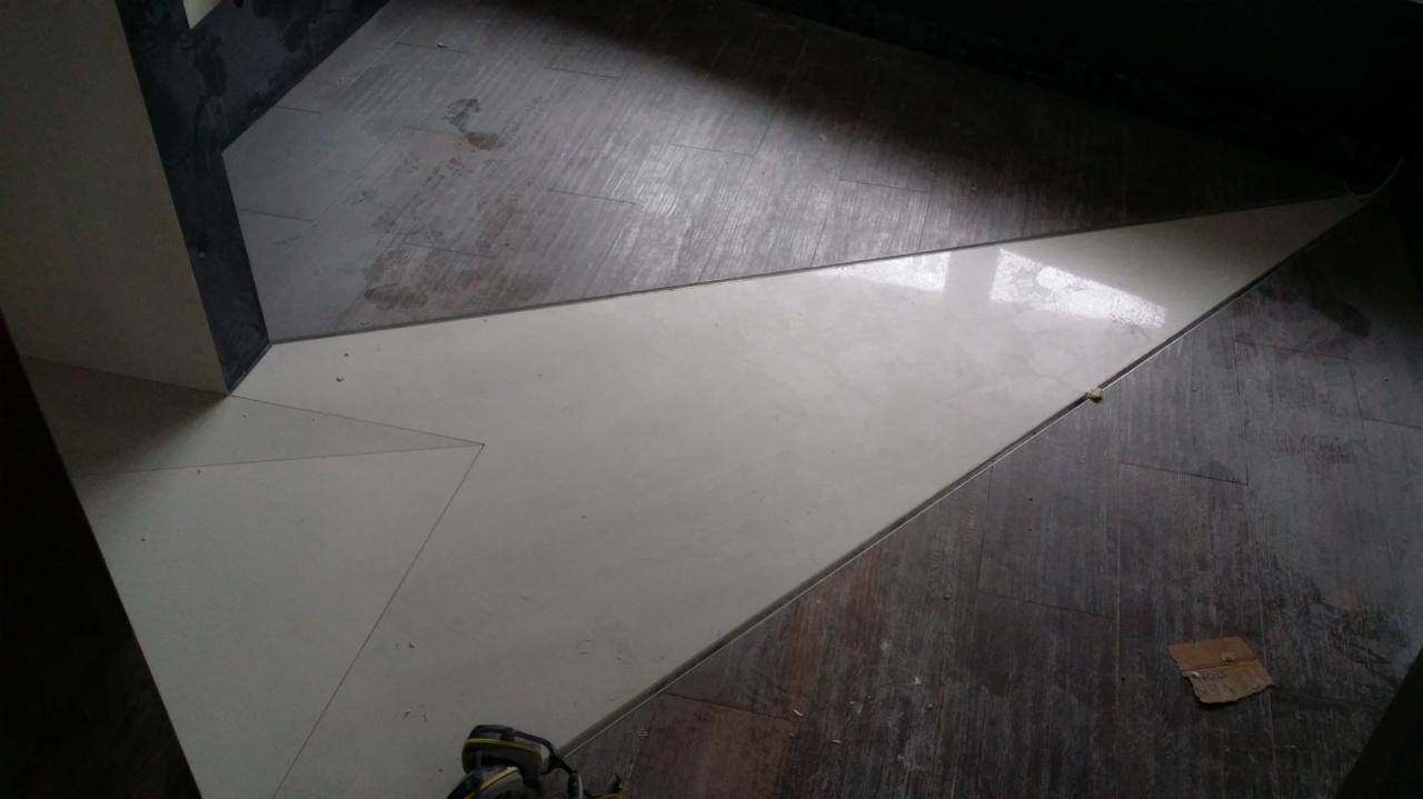 Ułożona podłoga z płytek w dwóch kolorach: jasny beż i ciemne drewno, ułożone w geometryczny wzór trójkątów, widoczne narzędzie do cięcia płytek w rogu.