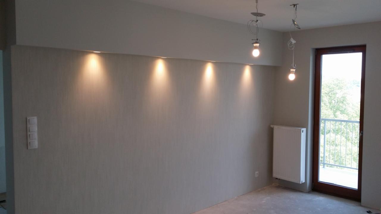 Szara ściana z ukrytymi punktowymi źródłami światła, surowe żarówki na kablach, biały grzejnik i okno balkonowe z widokiem na zieleń