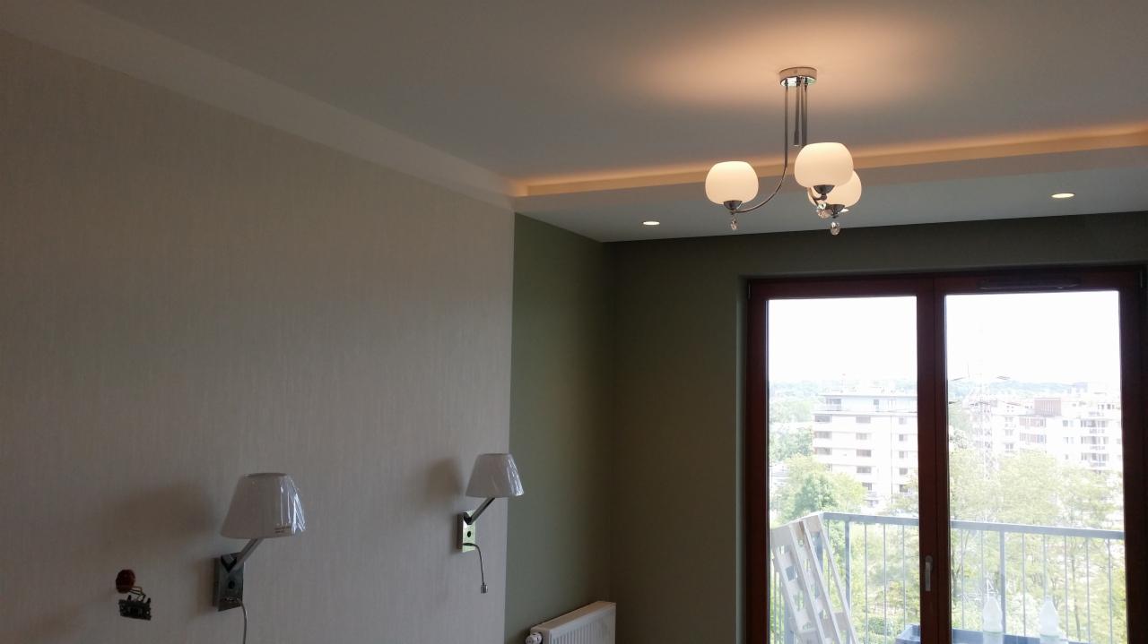 Pokój po remoncie: jasne ściany z tapetą, biały sufit z listwą, oświetlenie LED, lampa wisząca z trzema białymi kloszami, dwa kinkiety z białymi abażurami na ścianie, okno balkonowe z widokiem...