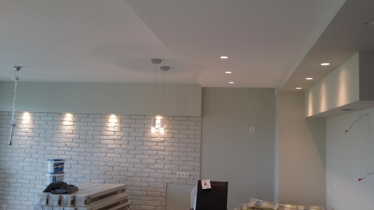 Wykończone wnętrze: biała ceglana ściana z oświetleniem punktowym i wiszącymi lampami, świeżo pomalowany sufit i ściany w odcieniach szarości, materiały budowlane w folii zabezpieczającej na podłodze.