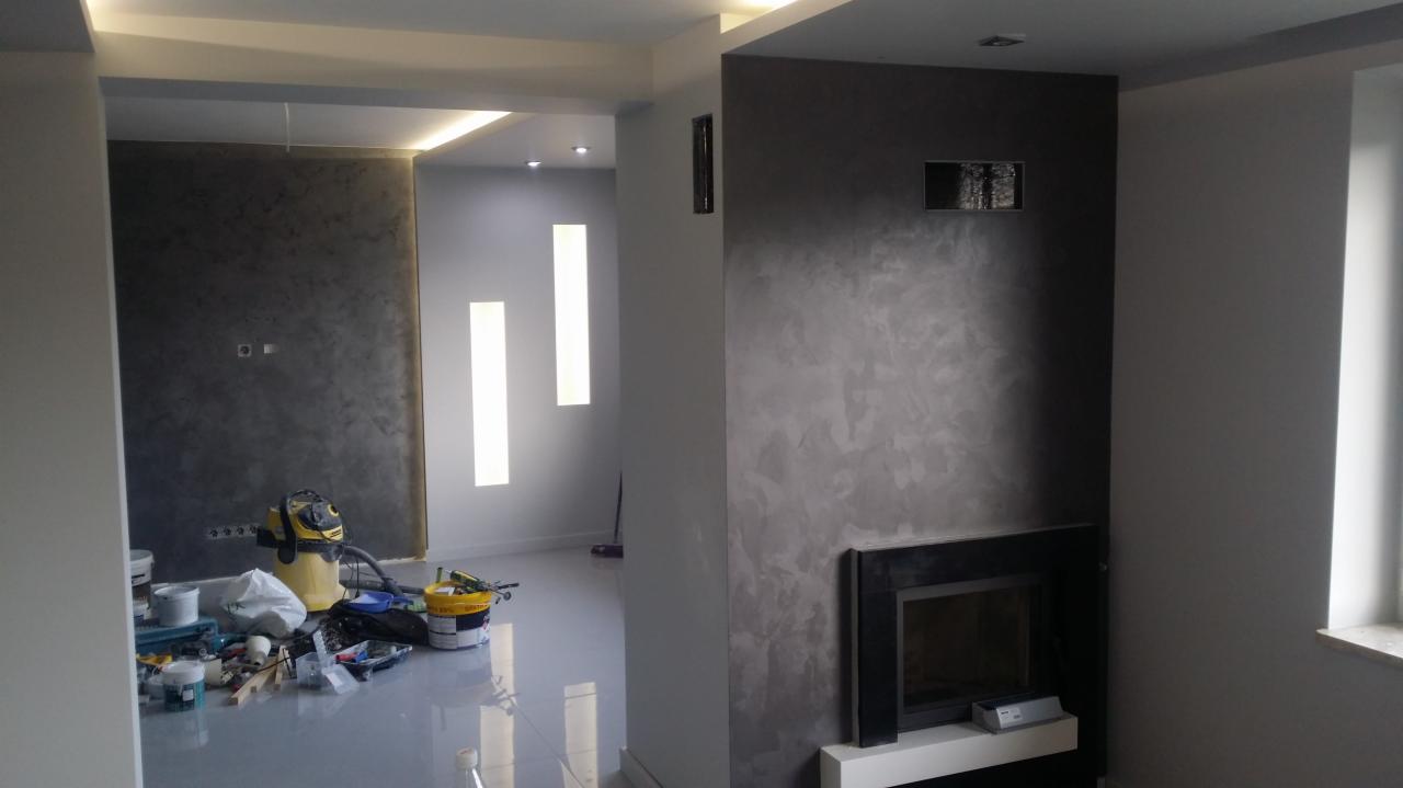 Pokój w trakcie prac wykończeniowych: ściany pokryte dekoracyjnym tynkiem w odcieniach szarości, widoczny kominek z czarną obudową, narzędzia i akcesoria malarskie rozłożone na podłodze, odkurzacz...